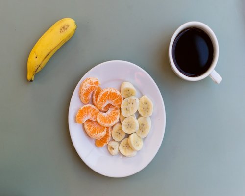 A freshly prepared single portion of sliced banana and organic fruits arranged on a plate next to morning hot tea